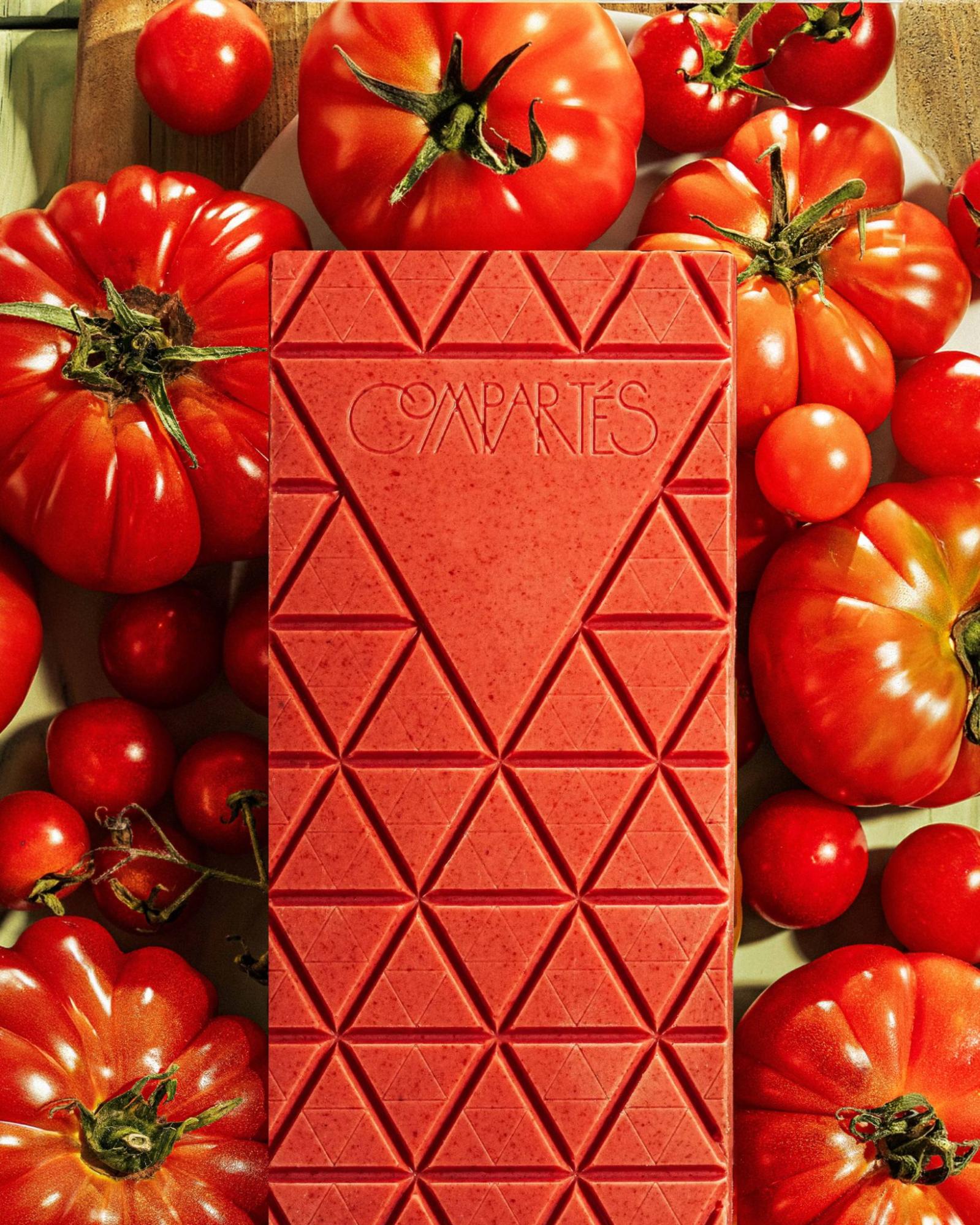 Compartés unusual tomato chocolate bar in vivid red with geometric triangle scoring, surrounded by fresh heirloom and cherry tomatoes — a bold and unexpected artisan chocolate flavor