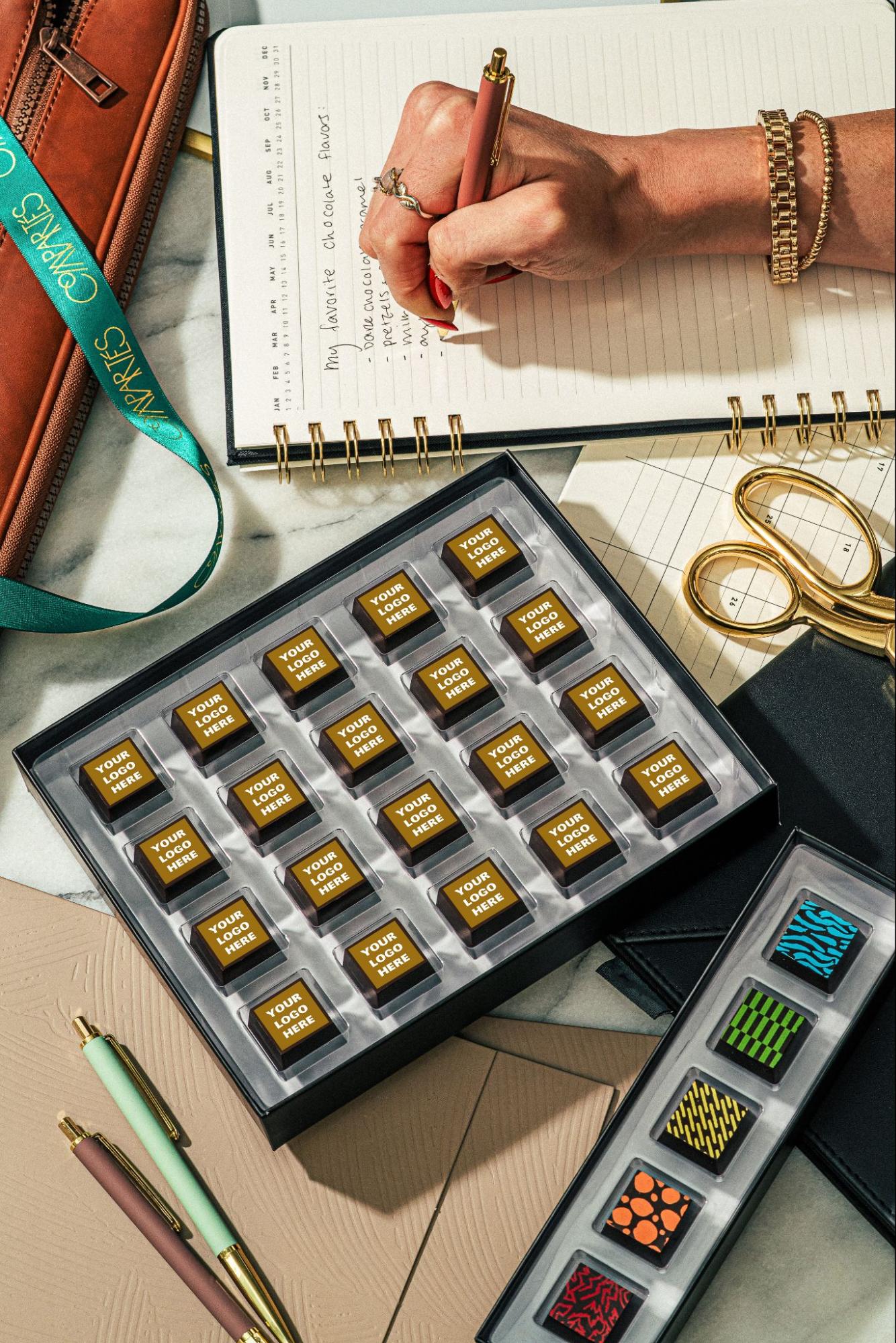 Compartés custom logo chocolates in a gift box with gold "Your Logo Here" squares as employee appreciation gifts, styled with a planner, gold scissors, pens, and a box of colorful patterned bonbons on a marble desk.