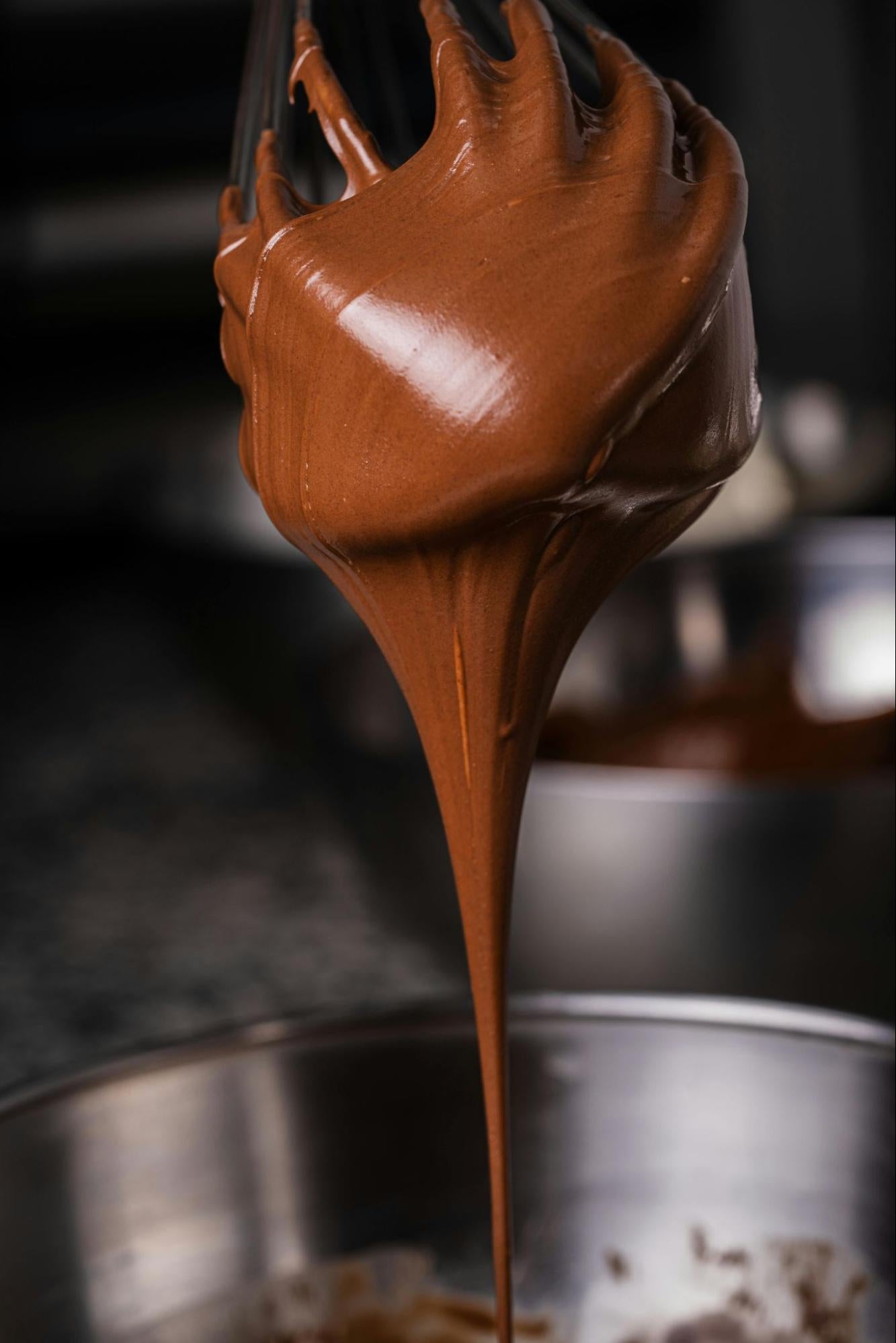 A close-up shot of thick, glossy milk chocolate mixture coating a wire whisk, with a long stream dripping down into a stainless steel mixing bowl.