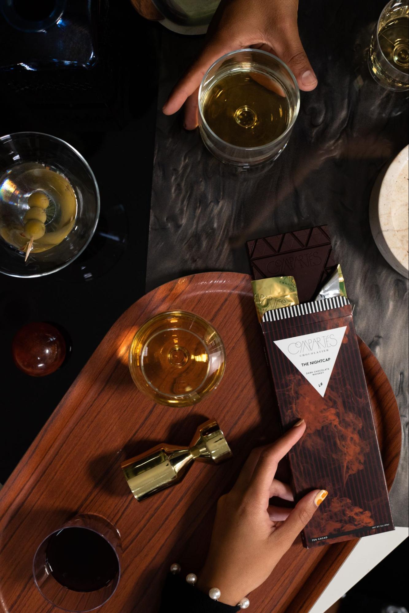 Hands holding Compartés 'The Nightcap' dark chocolate whiskey bar alongside cocktails and a gold jigger on a walnut tray — an elegant and unique chocolate anniversary gift for couples