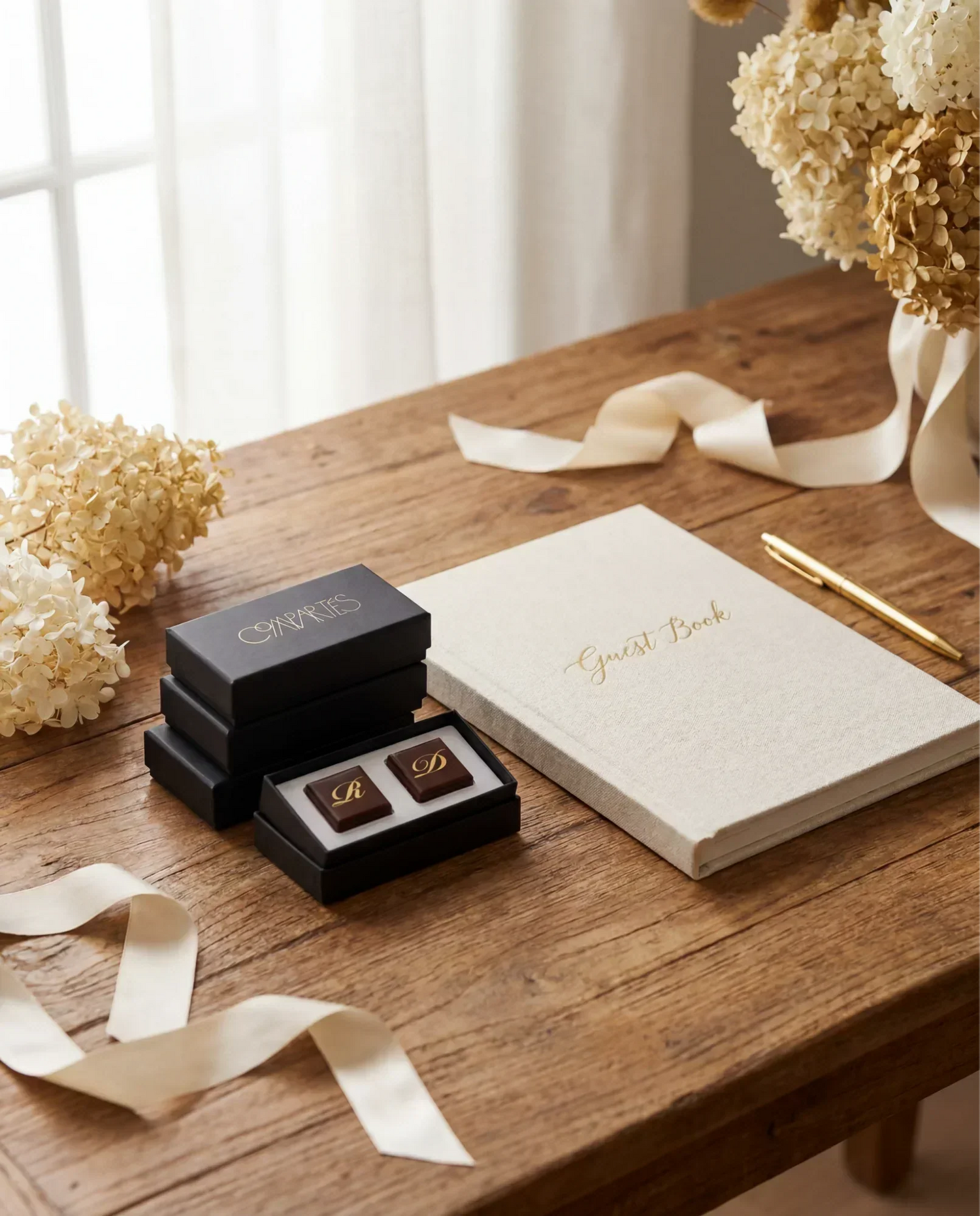 Compartés personalized wedding chocolates with gold monogram initials in an elegant black gift box, displayed beside a linen guest book and gold pen with dried hydrangeas as a keepsake wedding favor.