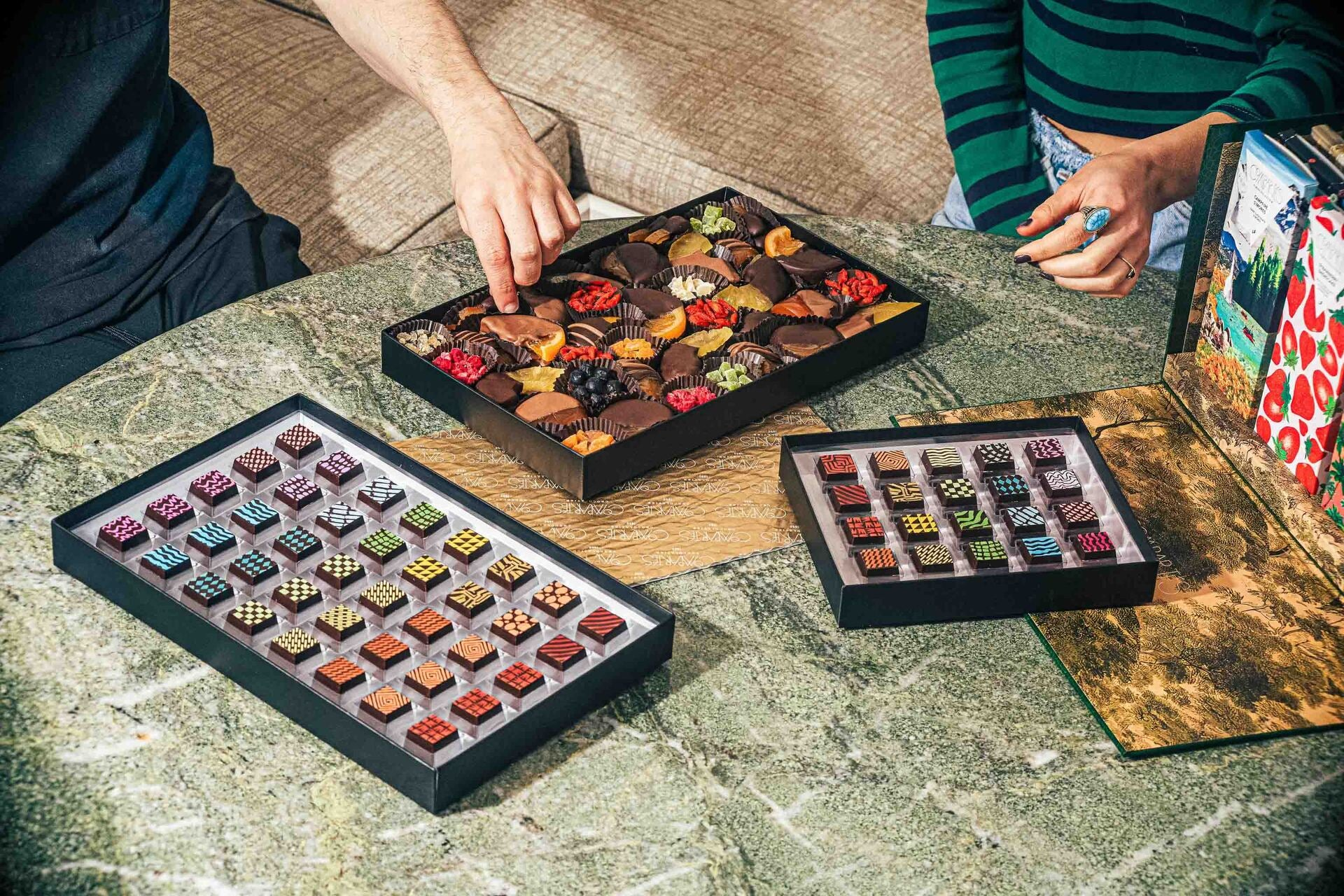 A high-angle shot shows two people sitting around a green marble table featuring three open boxes of Compartés gourmet chocolates.
