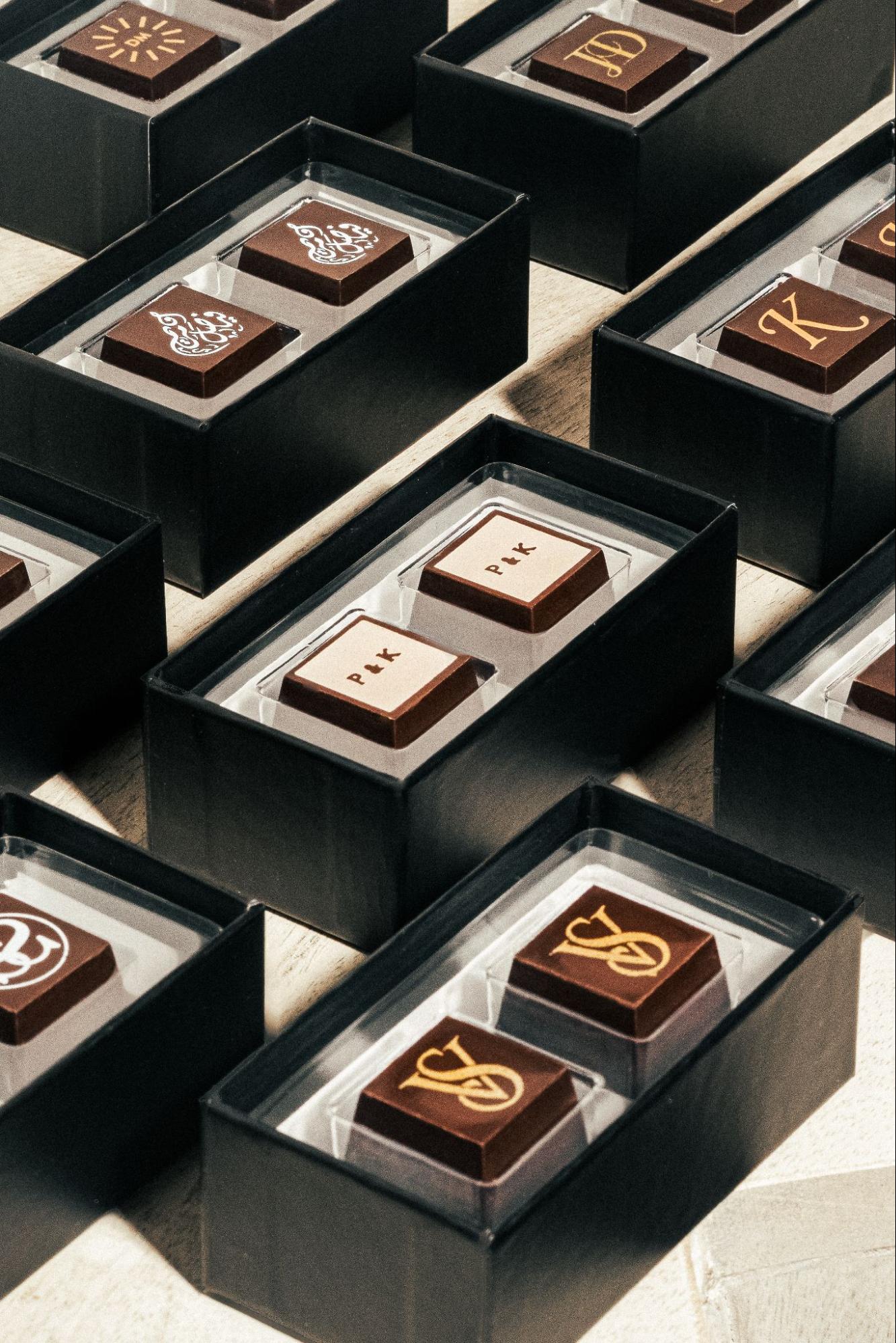 Custom monogrammed wedding favor chocolates in sleek black gift boxes, featuring personalized initials and elegant logo designs on artisan dark chocolate bonbons — a luxurious and unique wedding favor for guests