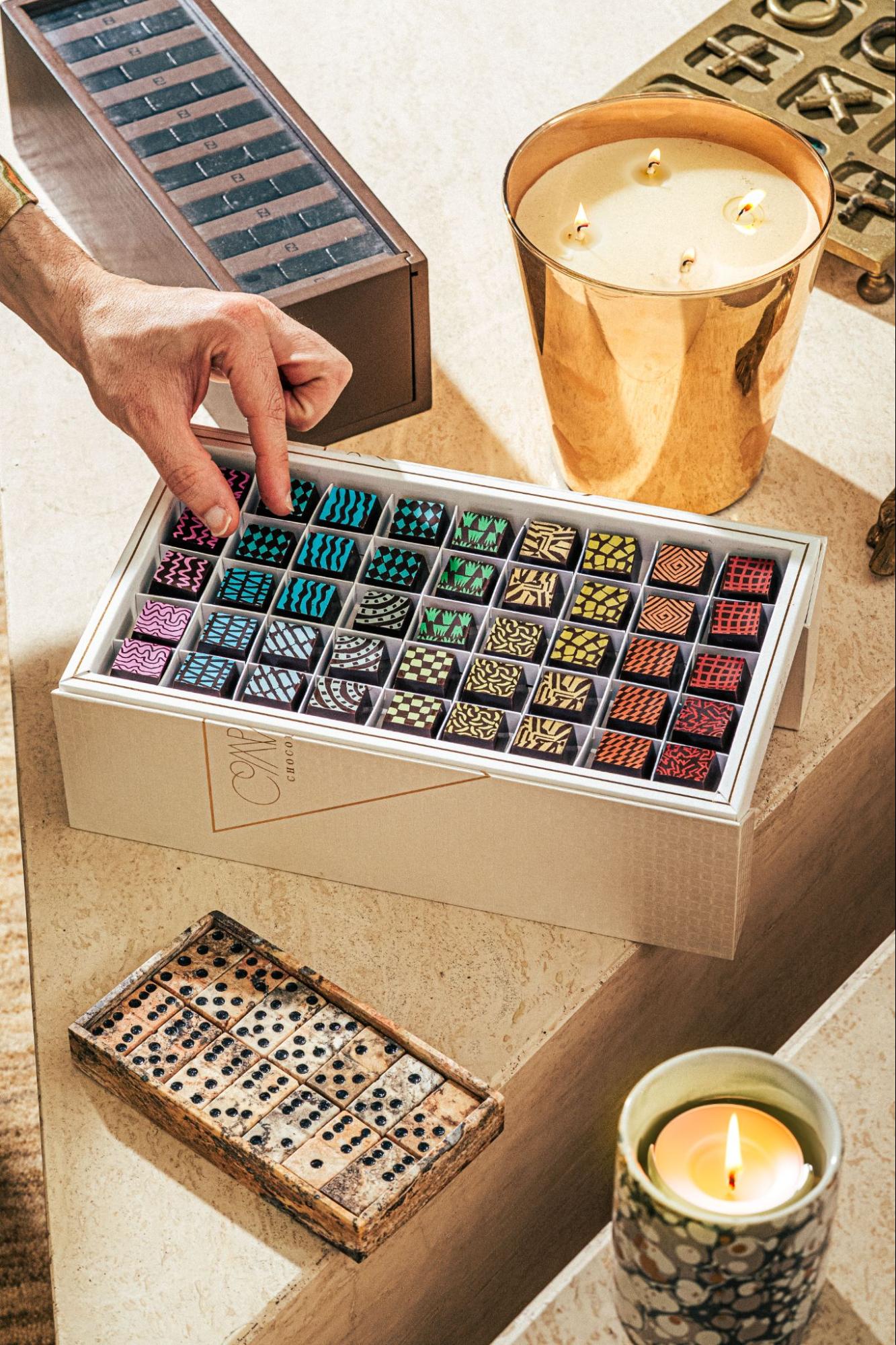 Hand selecting from a luxury box of the best chocolates, featuring artisan dark chocolate bonbons with colorful hand-painted geometric patterns in teal, pink, gold, and red, displayed in an elegant white gift box