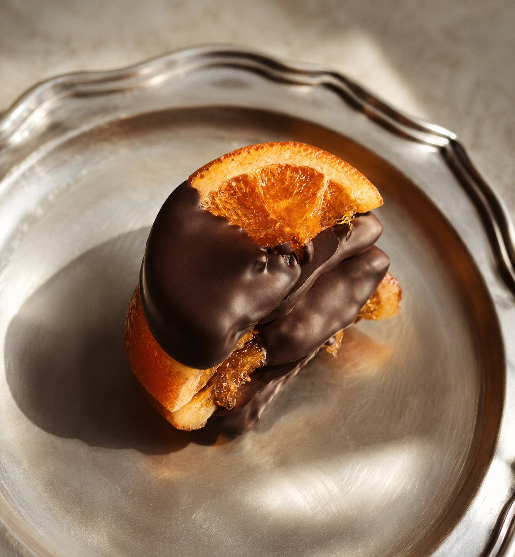 A close-up of a gourmet chocolate-covered orange, presented on a silver plate with dramatic sunlight and shadows.