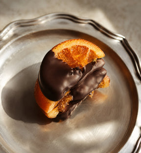 A close-up of a gourmet chocolate-covered orange, presented on a silver plate with dramatic sunlight and shadows.