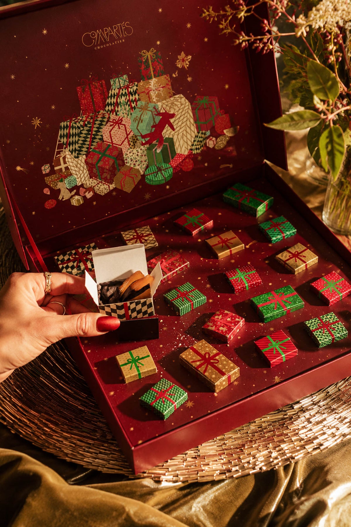 An open red Christmas-themed chocolate box with multiple wrapped chocolates arranged inside. Handmade gourmet luxury Christmas chocolates inside each little box.