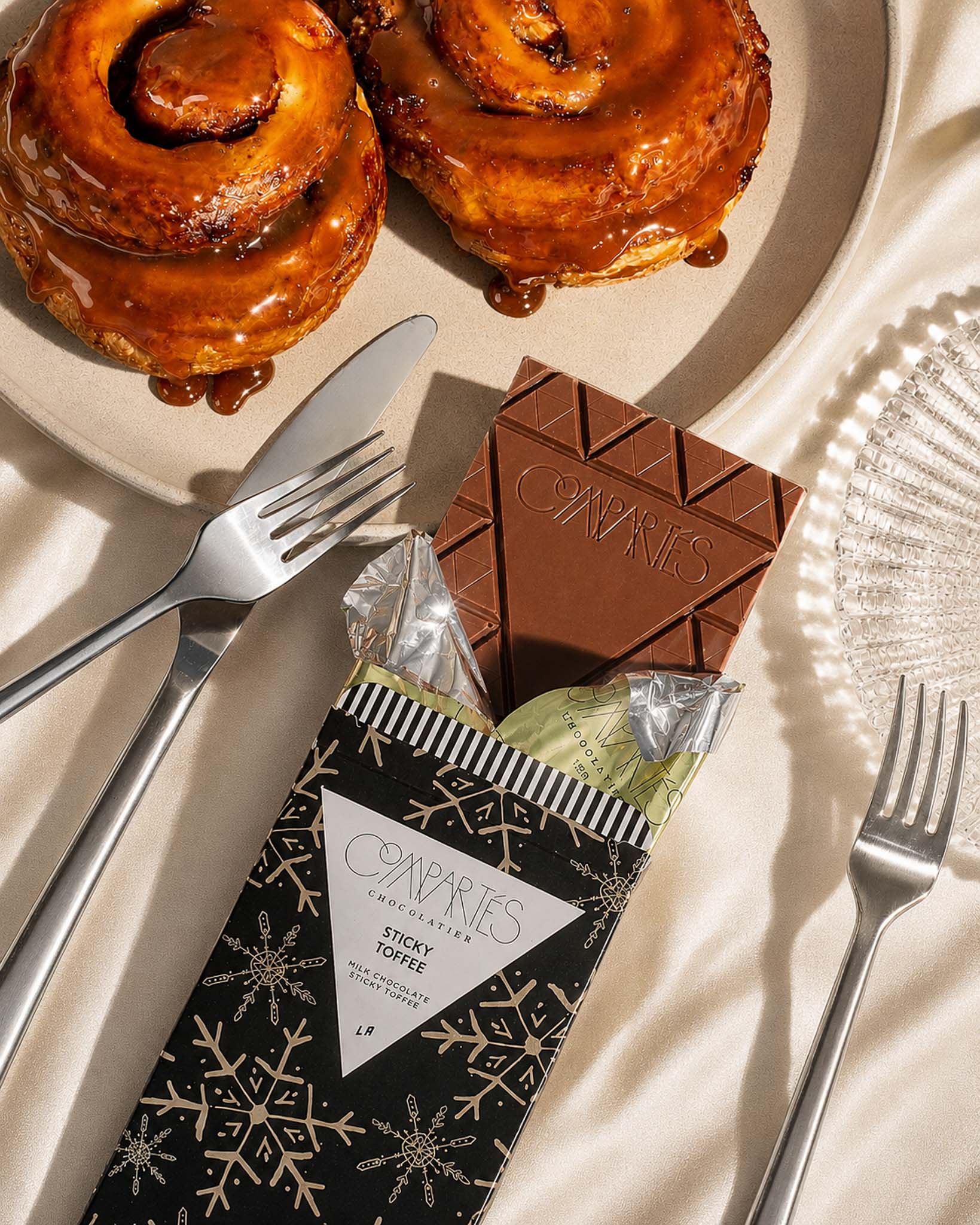 A packaged chocolate treat from Compartes Chocolate Bar, featuring a black box with gold snowflakes labeled 'Sticky Toffee' and a dark brown chocolate bar.