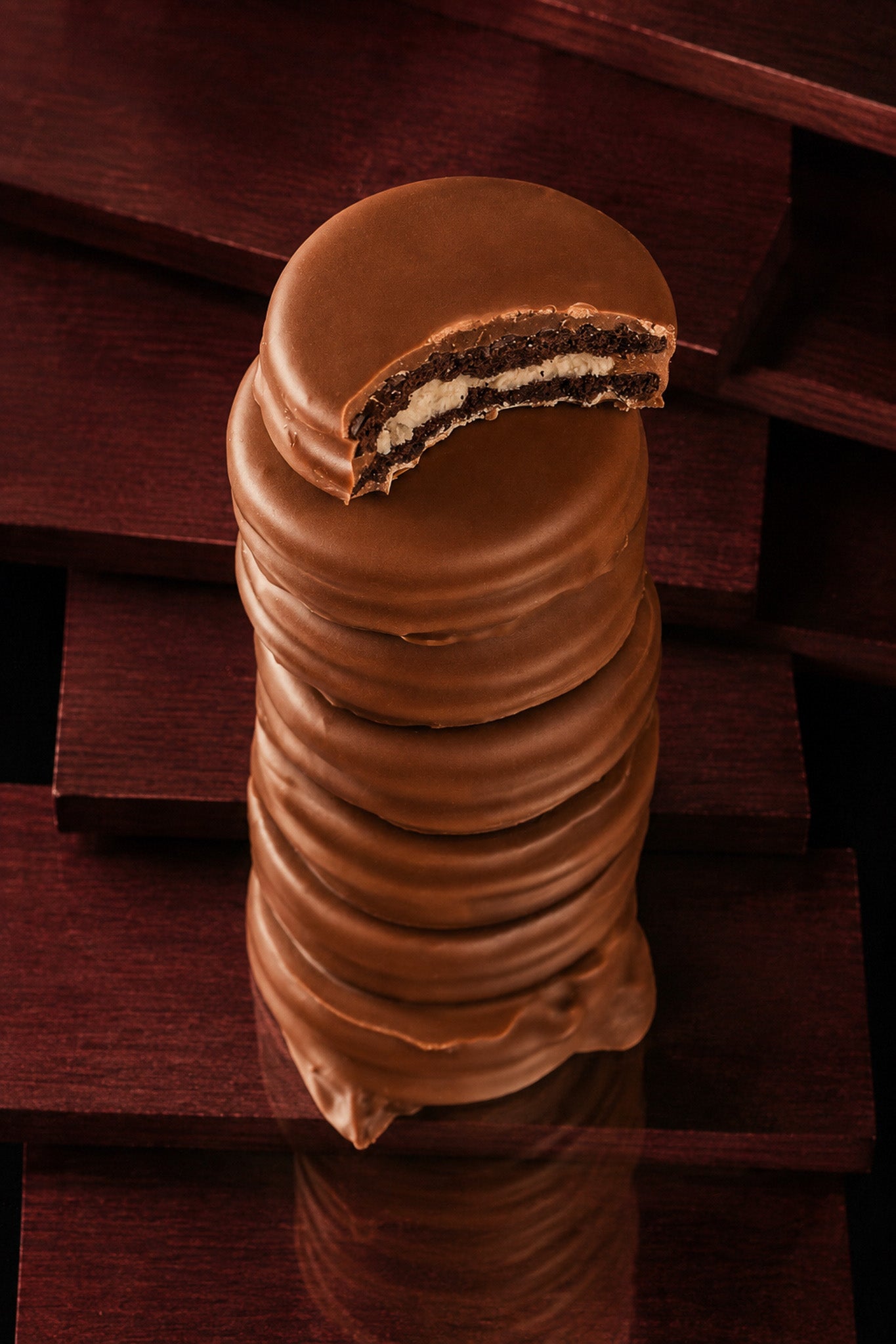 A stack of chocolate-covered Oreos cookies with a bite taken out at the top, showing a dark cookie and white cream filling, set against a dark wooden background.