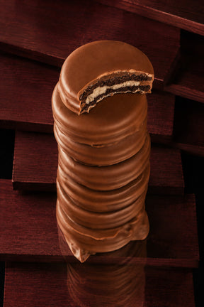 A stack of chocolate-covered Oreos cookies with a bite taken out at the top, showing a dark cookie and white cream filling, set against a dark wooden background.
