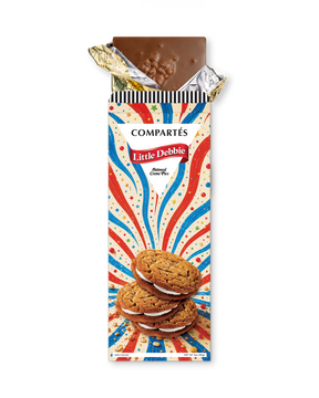 A Compérés Little Debbie oatmeal creme pies bar in its wrapper, with the top foil partially peeled back to reveal a piece of chocolate. The wrapper features a red, white, and blue burst design with an illustration of oatmeal creme pies.